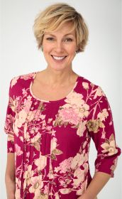 Frau mit kurzen blonden Haaren lächelt, trägt baumwoll-mix-kleid in Magenta mit floralem Muster in Rosa und Grün, 3/4-Arm, lockere Passform, helle Hintergrundfarbe