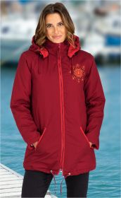 Frau trägt rote Baumwoll-Jacke mit Kapuze, Reißverschluss und eingesticktem Blumenmuster auf der Brust, Hände in den Taschen, steht vor unscharfem Wasser- und Bootshintergrund