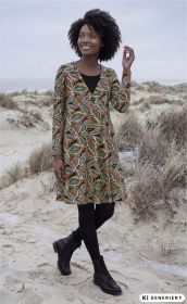 Frau am Strand trägt langärmliges Baumwoll-Mix-Kleid mit buntem, floralem Muster, schwarze Strumpfhose und schwarze Schnürstiefel, lächelt und blickt seitlich, sandige Dünen und lichte Vegetation im Hintergrund