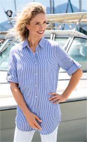 Frau mit kurzen blonden Haaren trägt lässige blau-weiß gestreifte Baumwoll-Bluse mit hochgekrempelten Ärmeln, steht vor einem Boot und lächelt, weiße Hose, sonniger Tag am Wasser