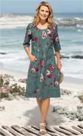 Frau am Strand trägt grünes Baumwoll-Kleid mit buntem Blumenmuster, 3/4-Ärmel, Taschen, weiße Sandalen, hält Strohhandtasche, steht auf Holzsteg mit Sand, Meer und Felsen im Hintergrund, sonniger Tag