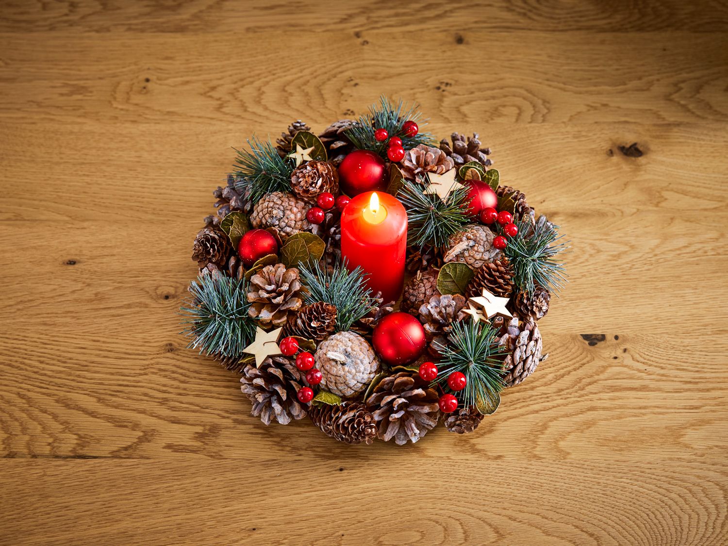 Deko-Kranz mit brennender roter Kerze, Tannenzapfen, roten Christbaumkugeln, Holzsternen, grünen Tannenzweigen und roten Beeren auf hellem Holztisch