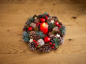 Deko-Kranz mit brennender roter Kerze, Tannenzapfen, roten Christbaumkugeln, Holzsternen, grünen Tannenzweigen und roten Beeren auf hellem Holztisch