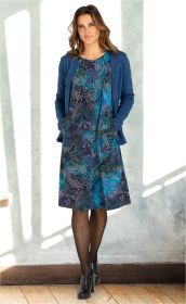 Frau trägt blau gemustertes Baumwoll-Mix-Kleid mit floral-abstraktem Muster, dazu eine blaue Strickjacke, schwarze Strumpfhose und schwarze Stiefeletten, steht vor hellem Hintergrund mit Holzfußboden