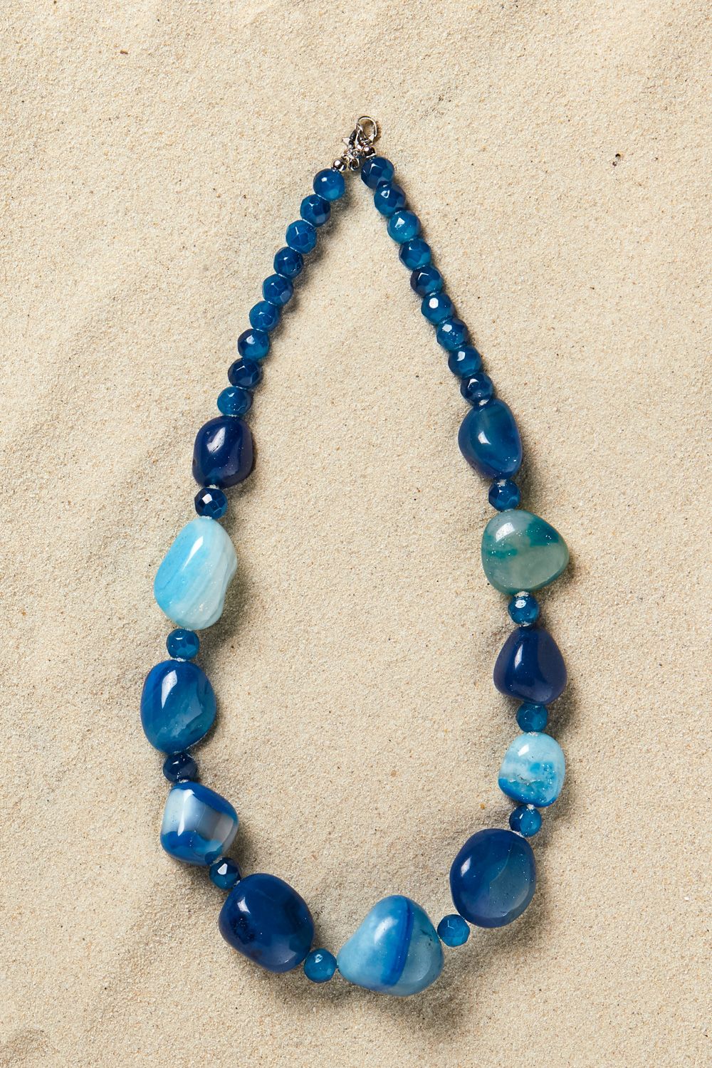 Achatkette ca. 48-50cm mit verschiedenen blauen, teils facettierten und glatten Achatperlen in unterschiedlichen Formen auf hellem Sandhintergrund.