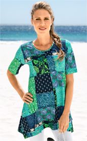 Frau am Strand trägt buntes Viskose-Mix-Shirt mit Patchwork-Muster in Blau-, Grün- und Türkistönen, kurze Ärmel, runder Ausschnitt, lockere Passform, helle Hose, Hand in Hüfte, sichtbarer Zopf, sonniger Tag