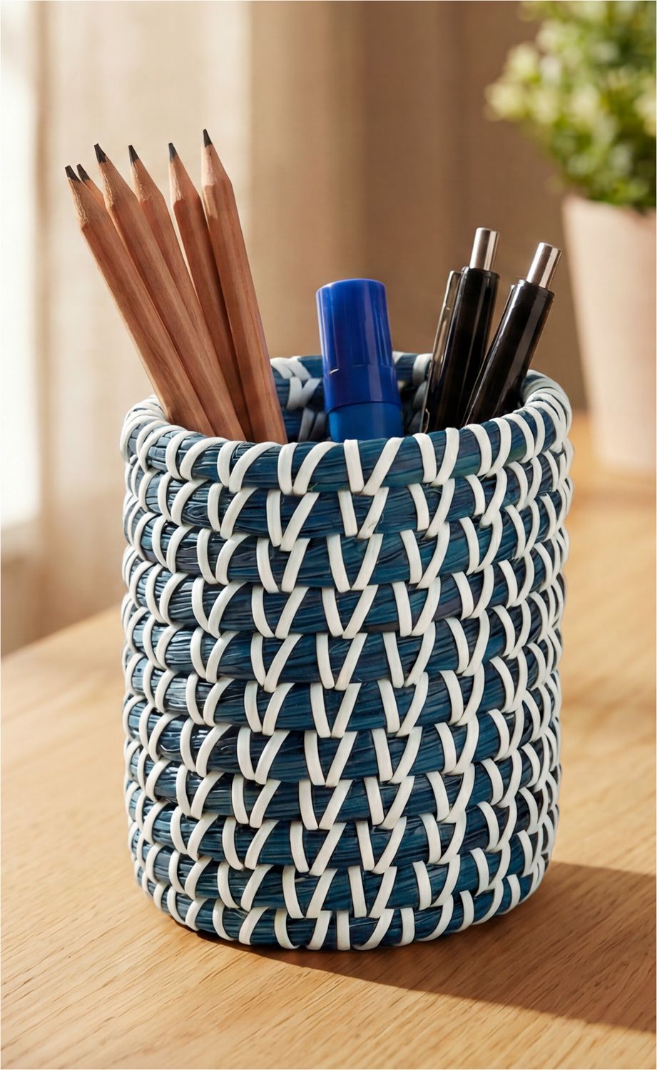 blauer geflochtener Stiftehalter mit weißen Akzenten auf Holztisch, enthält sechs Bleistifte, zwei schwarze Kugelschreiber und einen blauen Korrekturstift, unscharfer Hintergrund mit Pflanzenmotiv