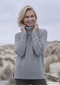 lächelnde blonde Frau mit kurzem Haar trägt grauen Rollkragenpullover am windigen Strand mit Dünen und trockenem Gras im Hintergrund