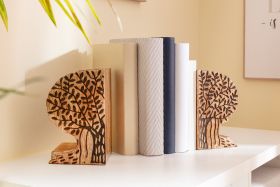 Set aus zwei hölzernen Buchstützen mit Baum-Motiv, hält mehrere Bücher mit neutralen Einbänden in beige, grau, schwarz und weiß auf einem weißen Tisch, helle Wand im Hintergrund, Pflanze oben links sichtbar