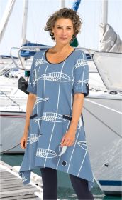 Frau trägt blaues Baumwoll-Kleid mit weißem Fisch- und Linienmuster, kurze Ärmel mit Umschlag, zwei dunkle Taschen, steht vor Segelbooten im Hafen, trägt dunkle Leggings