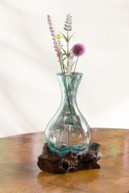 Glasvase mit bauchiger Form auf einem rustikalen Wurzelholzuntersetzer, gefüllt mit Trockenblumen in verschiedenen Farben und Formen, auf einem Holztisch vor heller Wand