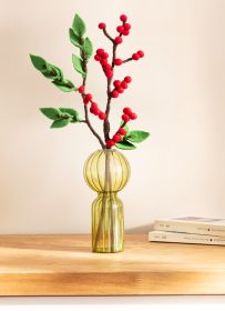 Zwei Filz-Zweige mit roten Beeren und grünen Blättern in einer gelben, gerippten Glasvase auf einem Holztisch, dahinter Stapel mit Büchern, heller Hintergrund