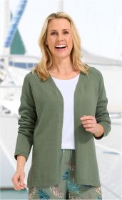 Frau mit blonden Haaren trägt offene, olivgrüne Baumwoll-Strickjacke über weißem Shirt, dazu gemusterte Hose mit Blattmotiven, lächelt freundlich, Hintergrund mit unscharfen Segelbooten und Wasser.