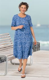Frau in blauem Viskose-Kleid mit weißem Blumenmuster, kurzen Ärmeln und rundem Ausschnitt, läuft barfuß mit weißen Flip-Flops auf Holzsteg am Meer, lächelt freundlich, im Hintergrund Wasser und verschwommener Segelboot.