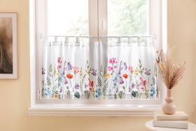 Weiße Küchengardine mit buntem Blumenmuster, hängend an einer Stange vor einem hellen Fenster, umgeben von beiger Wand, links ein gerahmtes Bild, rechts eine Vase mit Trockenblumen auf zwei Büchern aufgeräumt auf einem kleinen runden Tisch