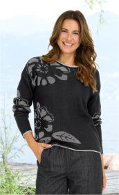 lächelnde Frau mit braunen welligen Haaren, trägt schwarzen Pullover mit großen grauen Blumenmustern, Hand in der Hosentasche, sitzende Kulisse mit Wasser und Pflanzen im Hintergrund