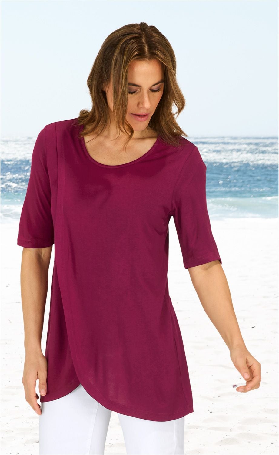 Frau trägt legeres, kastanienbraunes Viskose-Shirt mit Dreiviertelärmeln und rundem Ausschnitt, am Strand mit Meer im Hintergrund, kombiniert mit weißen Hosen.
