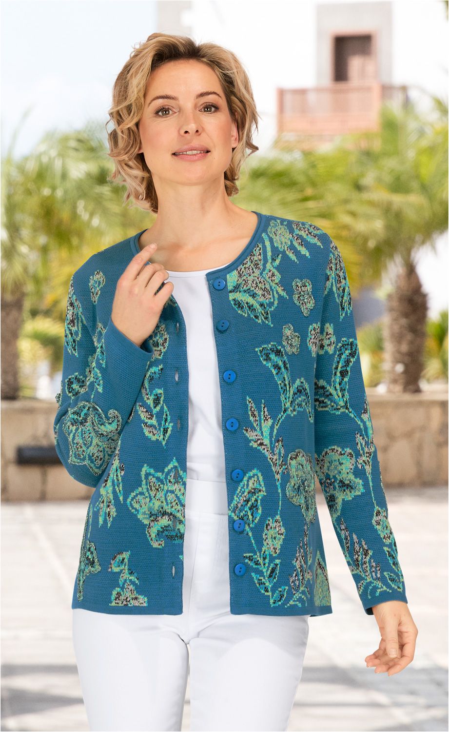 Frau trägt blaue Baumwoll-Strickjacke mit grünem Blumenmuster, Knopfleiste, darunter weißes Shirt und weiße Hose, steht draußen bei Sonnenschein mit verschwommenen Palmen im Hintergrund