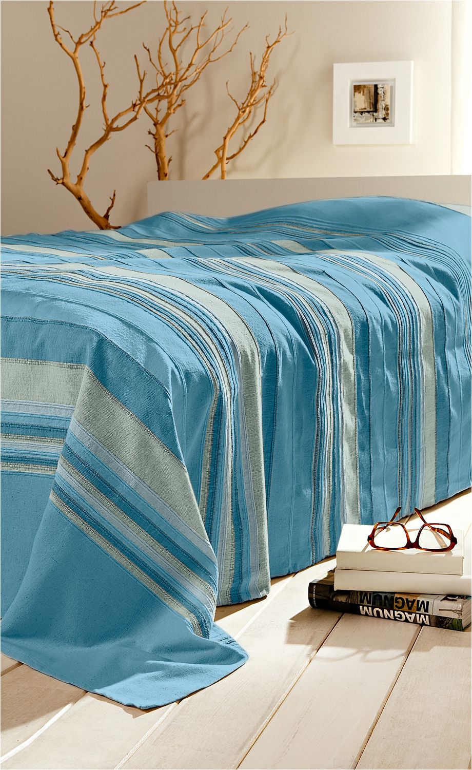 Baumwoll-Überwurf 250x180 in Blau mit gestreiftem Muster auf hellem Holzboden, dekorativ drapiert, im Hintergrund weiße Wand mit abstraktem Bild und Zweigen, neben dem Überwurf Bücherstapel mit brauner Brille darauf