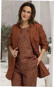 Frau trägt rostfarbene Leinen-Bluse mit 3/4-Ärmeln, dazu gestreiftes, rostfarbenes Oberteil und Hose, steht vor hellem Hintergrund mit Pflanzen, lächelt, rechte Hand in Hosentasche, linke Hand hält Bluse geöffnet