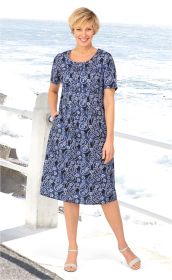 Frau in blauem Baumwoll-Mix-Kleid mit weißem Paisley-Muster, kurzen Ärmeln, Stehkragen und Taschen, steht lächelnd am Strand mit Meer im Hintergrund, trägt weiße Sandalen