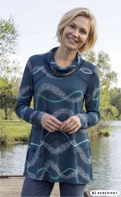 Frau mit kurzem blondem Haar trägt ein blau-grünes, langärmeliges Viskose-Shirt mit Muster aus weißen und türkisfarbenen Kreisen und Linien, steht lächelnd am Wasser mit Bäumen im Hintergrund