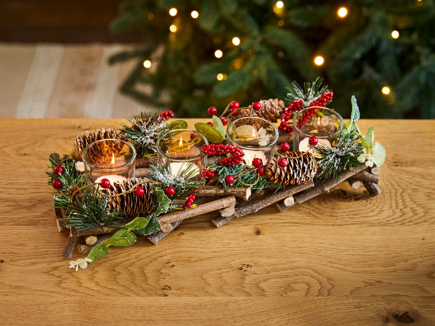 Teelichthalter aus Holzstäben, dekoriert mit roten Beeren, Tannenzapfen, grünen Zweigen und weißen Blüten, vier brennende Teelichter in Glasbehältern, warmes Licht, weihnachtliche Tischdekoration auf Holzoberfläche