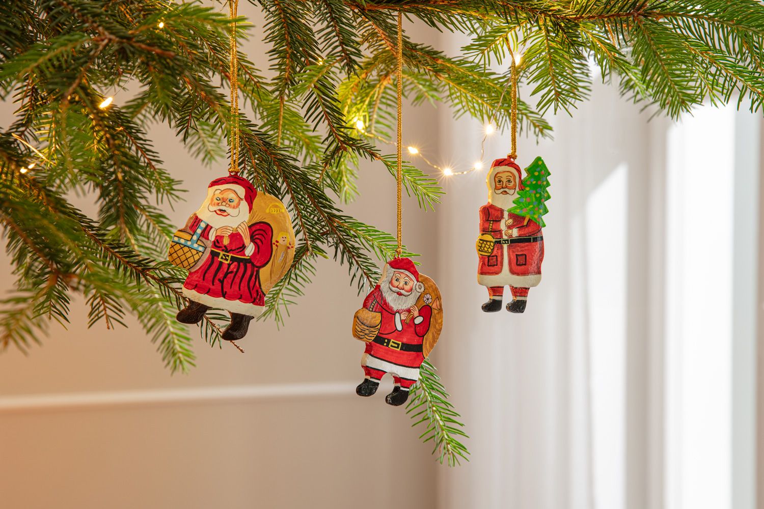 Drei handbemalte Weihnachtsmannhänger aus Holz mit Geschenksack, Lebkuchen und Tanne, hängend an grünen Tannenzweigen mit Lichterkette vor hellem Hintergrund