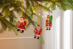 Drei handbemalte Weihnachtsmannhänger aus Holz mit Geschenksack, Lebkuchen und Tanne, hängend an grünen Tannenzweigen mit Lichterkette vor hellem Hintergrund