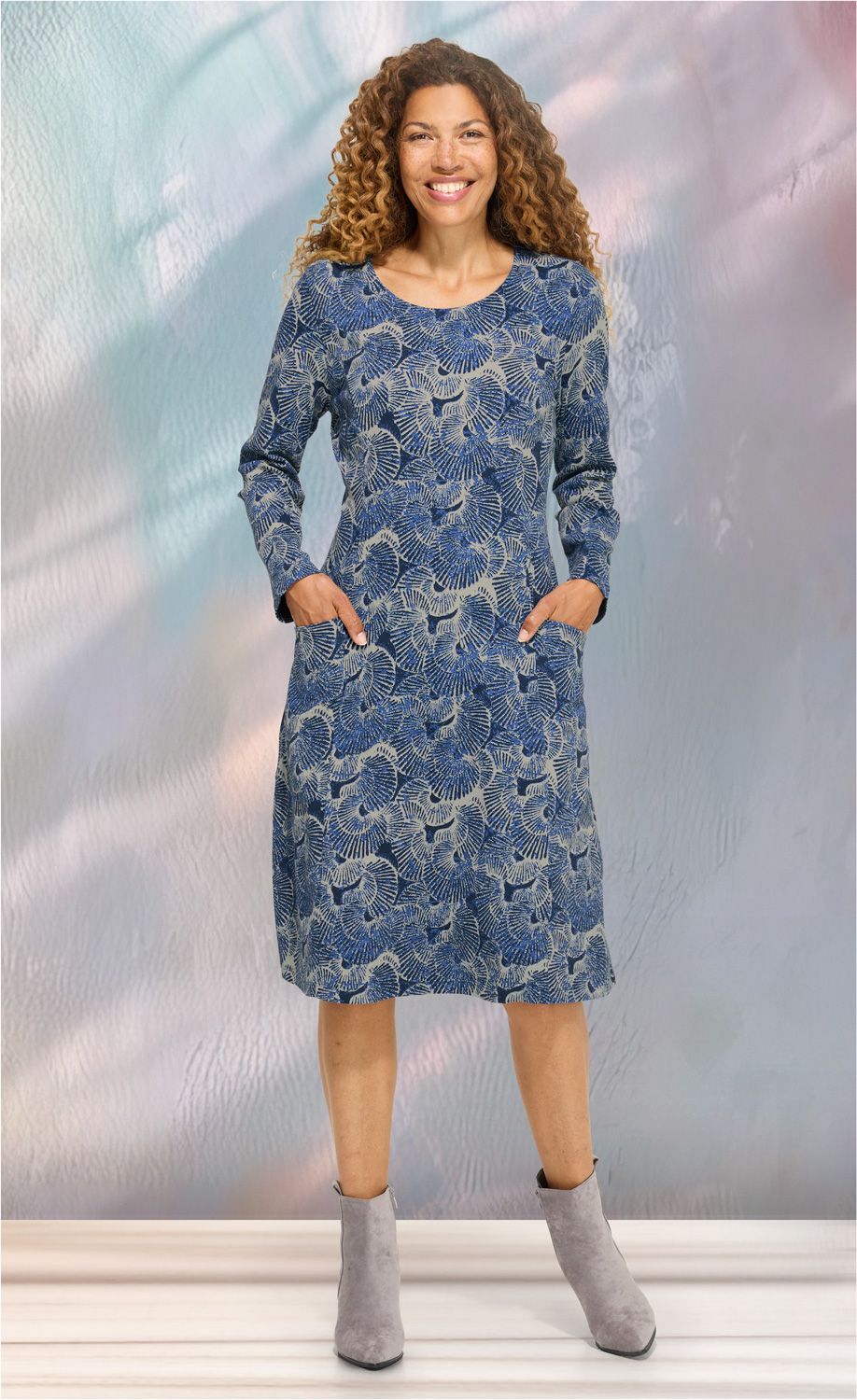 Frau mit lockigen Haaren trägt blau-beiges Baumwoll-Kleid mit floralem Muster, langen Ärmeln und Taschen, graue Stiefeletten, steht vor pastellfarbenem Hintergrund, lächelt, Ganzkörperaufnahme