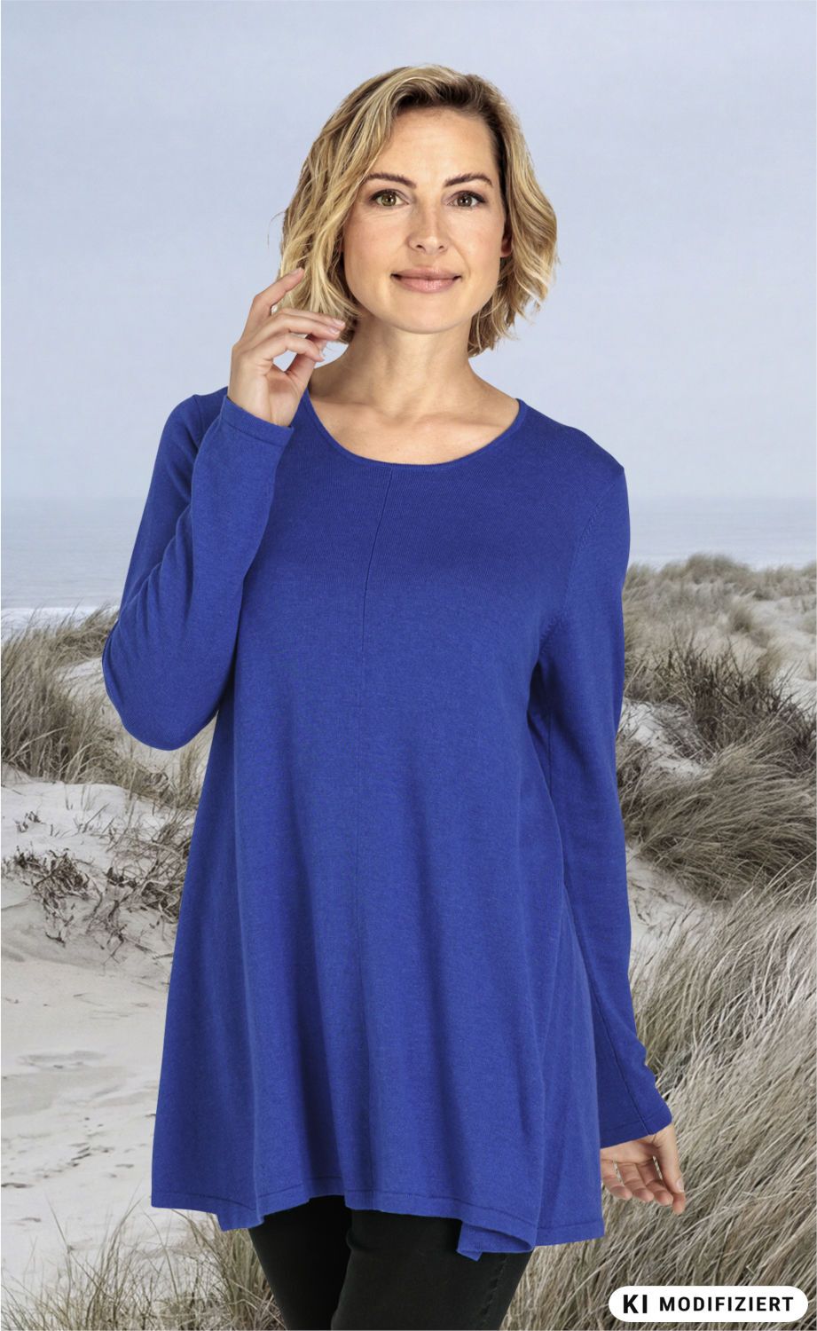 Frau mit blondem, schulterlangem Haar trägt blauen Viskose-Mix-Pullover mit Rundhalsausschnitt, steht vor sandiger Dünenlandschaft mit Strandgras, Blick leicht lächelnd, eine Hand am Gesicht, schwarze Hose sichtbar