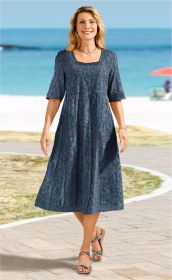 Frau am Strand trägt ein mittellanges dunkelblaues Baumwoll-Kleid mit halbtransparentem Blumenmuster, halblangen Ärmeln und quadratischem Ausschnitt, kombiniert mit bunten Sandalen, lächelnd, blonder schulterlanger Haarstil, sommerliche Strandkulisse.
