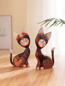 Zwei farbenfrohe, handbemalte Deko-Katzen aus Holz mit blau betonten Augen, roten und weißen Akzenten und unterschiedlichen Mustern, sitzend auf hellem Holztisch vor hellem, wohnlichem Hintergrund mit Pflanzenblättern.