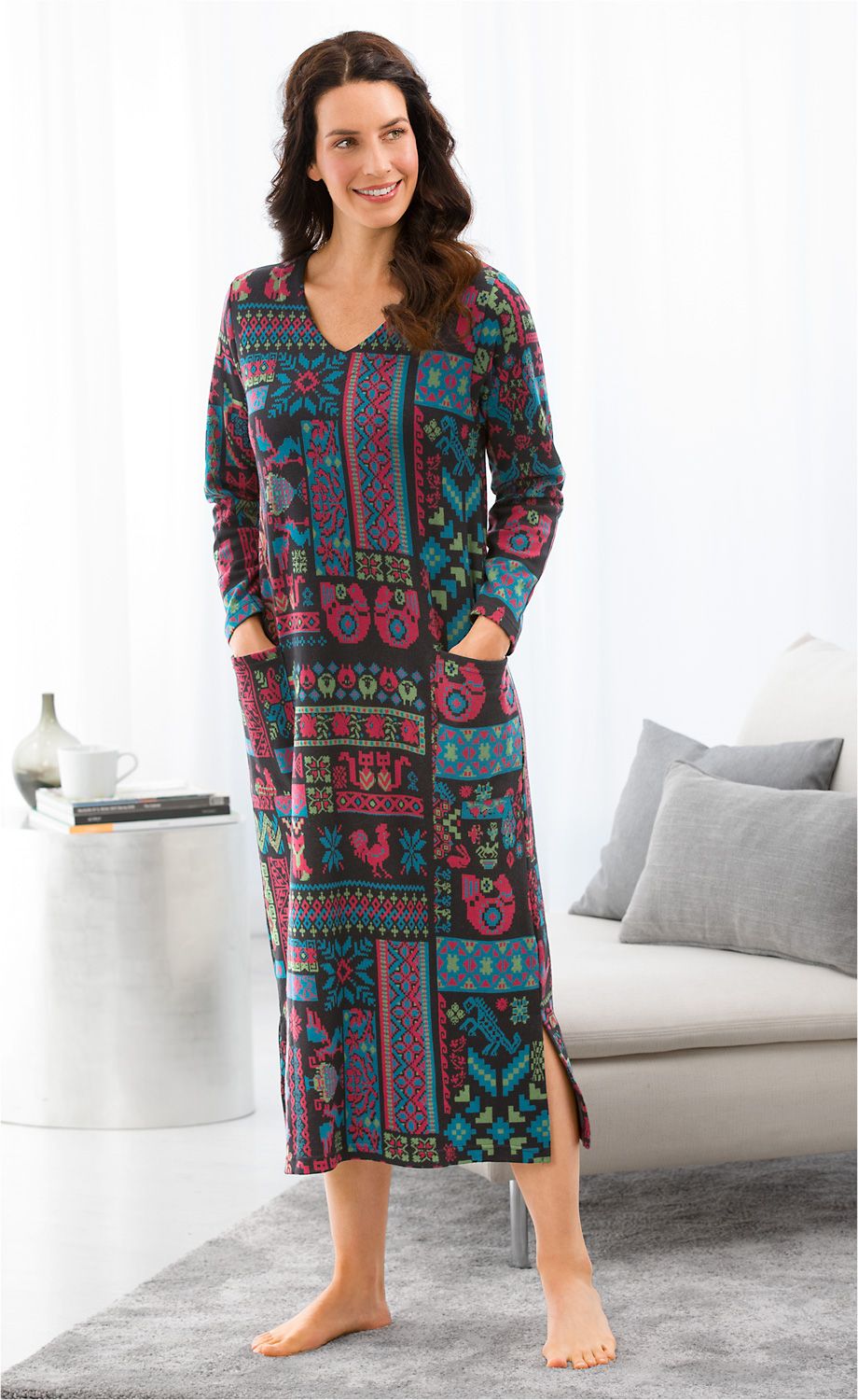 Frau trägt farbenfrohes, langärmeliges Baumwoll-Hauskleid mit geometrischem und floralen Muster, seitlicher Schlitz, steht barfuß im Wohnzimmer, Hände in den Taschen, lächelt, helle Gardinen und graues Sofa im Hintergrund.