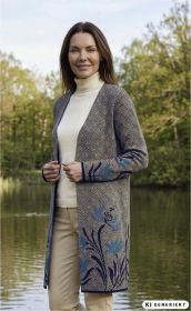 Frau trägt beige Baumwoll-Strickmantel mit geometrischem Muster und blauen Blumen, kombiniert mit weißem Rollkragenpullover und beigen Hosen, stehend vor einem See und grünen Bäumen