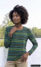 Frau trägt langärmliges Baumwoll-Shirt mit buntem, gestreiftem Muster in Blau-, Grün- und Gelbtönen, steht vor Fluss und Grünfläche, Hand berührt Kragen, lächelnder Ausdruck, beige Hose sichtbar.