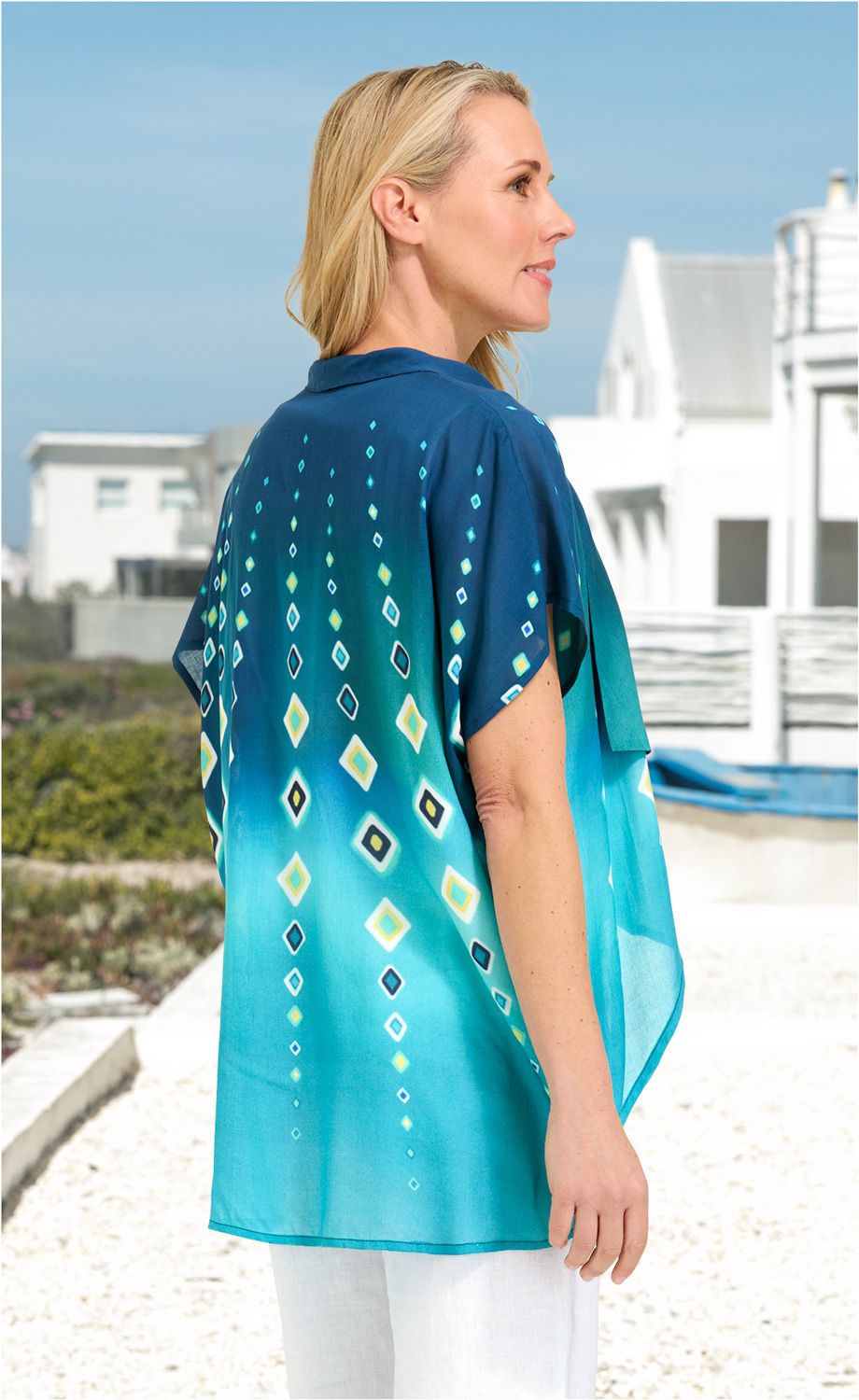 Frau von der Seite, trägt leichte Viskose-Bluse mit blau-grünem Farbverlauf und geometrischem Muster, weiße Hose, sommerliche Outdoor-Umgebung mit Häusern und klarem Himmel im Hintergrund