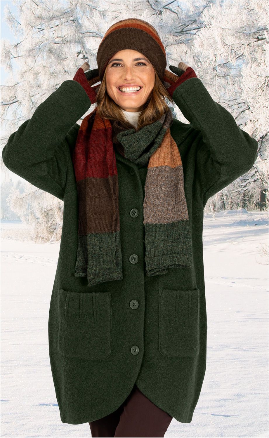 Frau trägt dunkelgrüne Woll-Jacke mit Knöpfen und aufgesetzten Taschen, bunte gestreifte Wollmütze, passende Wollhandschuhe und Schal, lächelt, steht vor verschneiter Winterlandschaft mit schneebedeckten Bäumen.