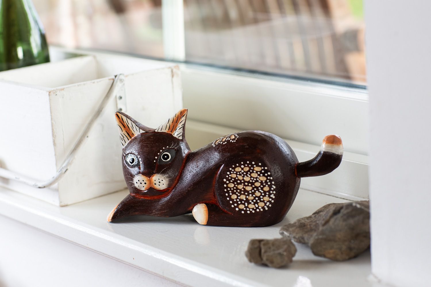 braune Deko-Katzenfigur aus Holz mit weißen Punkten und schwarzen Details, liegend auf weißem Fensterbrett neben weißem Holzkasten und Steinen, im Hintergrund unscharfes Fenster und Gartenbereich