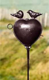 schwarzes Metallvogelbad in Herzform auf Stab mit zwei Vogelfiguren oben, unscharfer grüner Hintergrund