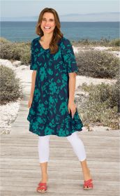 Frau trägt blaues Baumwoll-Mix-Kleid mit grünem Blumenmuster, weiße Capri-Leggings und rosa Sandalen, steht auf Holzsteg am Strand mit Meer und blühendem Küstengebüsch im Hintergrund, lächelt direkt in die Kamera