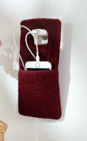 Burgunderrote, filzartige Handyladetasche, aufgehängt an Steckdose, darin ein weißes Smartphone mit angeschlossenem Ladekabel, das in einen weißen Netzstecker eingesteckt ist, weißer Hintergrund, Schatten an der Wand.