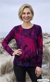 Frau mit blondem Bobhaar trägt langärmliges Baumwoll-Shirt in abstraktem Muster aus Pink, Violett und Schwarz, steht am Strand mit Dünen und Gras im Hintergrund, lächelt und hat eine Hand in die Hüfte gestützt.