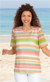 Frau am Strand trägt bunt gestreiftes Viskose-Mix-Shirt mit kurzen Ärmeln und Rundhalsausschnitt, lächelt, lockige braune Haare, weißer Unterteil, blauer Himmel und Meer im Hintergrund