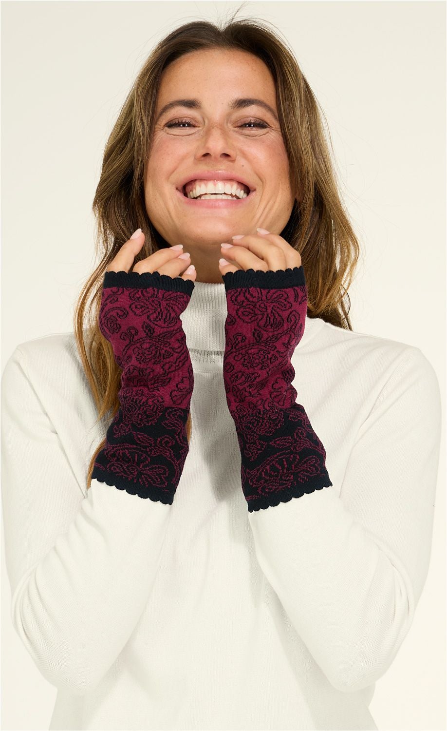 lächelnde Frau trägt weiße Rollkragenpullover und bordeauxrote Baumwoll-Pulswärmer mit schwarzem Blumenmuster, Finger frei und mit gewelltem Saum