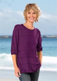 Frau am Strand trägt lächelnd violetten Leinen-Mix-Pullover mit halblangen Ärmeln und Rundhalsausschnitt, kombiniert mit blauen Jeans, im Hintergrund Meer und Himmel bei Tageslicht