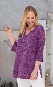 Frau trägt violette Viskose-Tunika mit rot-orangefarbenem Muster, 3/4-Ärmeln, Knopfleiste am Ausschnitt, kombiniert mit weißen Hosen, vor grauer Wand mit Tür