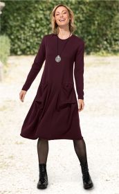 Frau trägt weinrotes, langärmeliges Baumwoll-Mix-Kleid mit runden Taschen, schwarze Netzstrumpfhose, schwarze Schnürstiefel und silberne Spiral-Kette, steht draußen vor grünem Hintergrund und lächelt.
