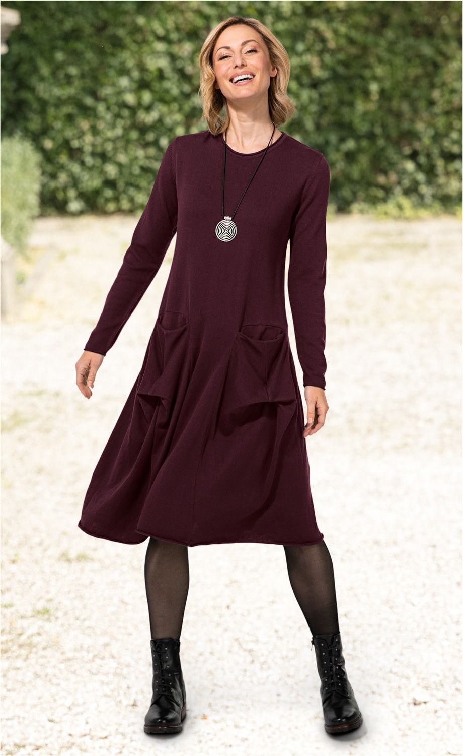 Frau trägt weinrotes, langärmeliges Baumwoll-Mix-Kleid mit runden Taschen, schwarze Netzstrumpfhose, schwarze Schnürstiefel und silberne Spiral-Kette, steht draußen vor grünem Hintergrund und lächelt.