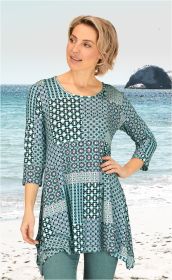Frau am Strand trägt Viskose-Mix-Shirt mit dreiviertel Ärmeln und blau-grünem Patchwork-Muster, dazu grüne Hose, Blick nach links, Hand in Hüfthöhe, sandiger Strand und Felsformation im Hintergrund